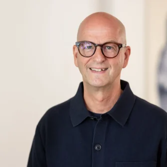 Lächelnder Mann mit Brille vor hellem Hintergrund.
